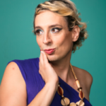 Grace Lavery wearing a blue shirt with blonde hair pulled back. Grace faces slightly to the left and has her hand raised to her chin, while making a slightly pouting expression.